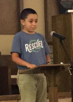 Young boy giving an LTC speech at a church lectern while wearing a “Rescued” T-shirt.