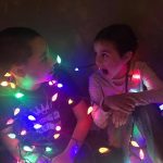 Two siblings playfully wrapped in colorful Christmas lights in 2021, continuing a family holiday tradition that began while living in Germany.