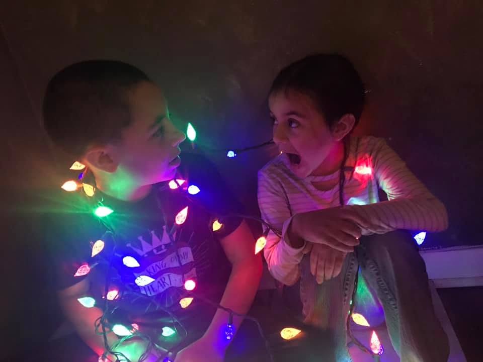 Two siblings playfully wrapped in colorful Christmas lights in 2021, continuing a family holiday tradition that began while living in Germany.