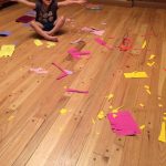 A young girl sits smiling in the middle of colorful paper scraps scattered across the floor during a craft project.