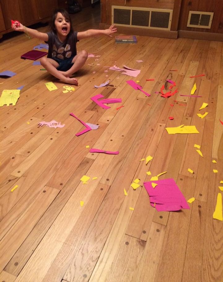 A young girl sits smiling in the middle of colorful paper scraps scattered across the floor during a craft project.