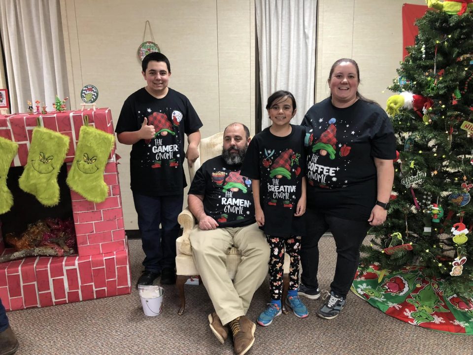 amily of four wearing matching Christmas gnome shirts beside a Grinch-themed Christmas tree and fireplace.