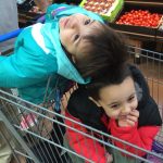 Two young children playfully pile together in a grocery cart during a family shopping trip