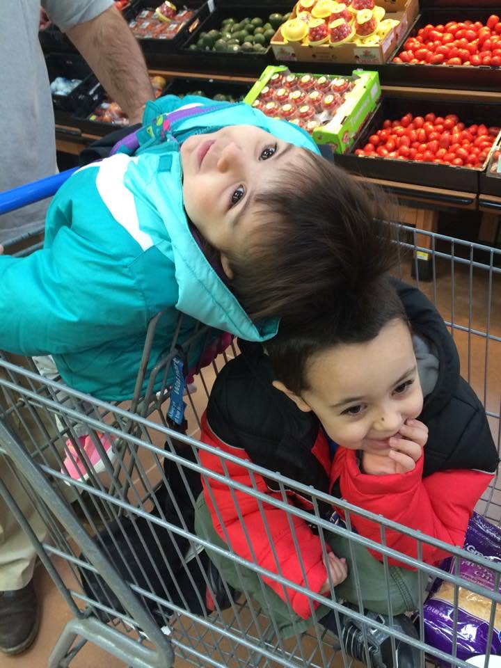 Two young children playfully pile together in a grocery cart during a family shopping trip