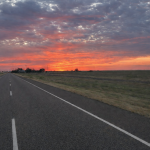 Straight two-lane road extending toward the horizon beneath a colorful sunset sky.