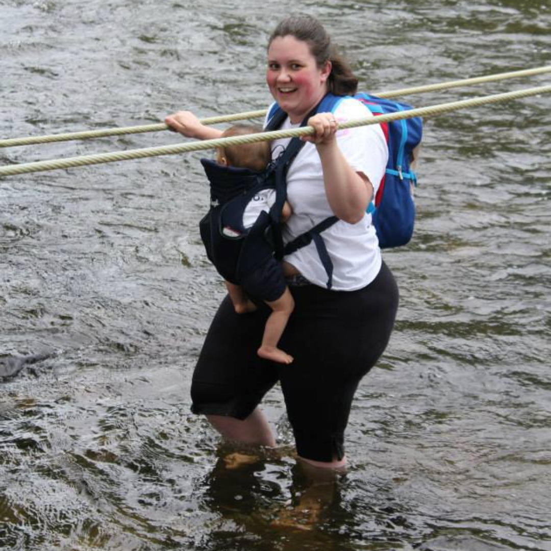 Angie walks through a shallow river carrying her young daughter in a front carrier while holding a rope for support at a barefoot park in Germany.