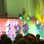Acappella performing on stage during a concert, with four singers visible in front of an audience.
