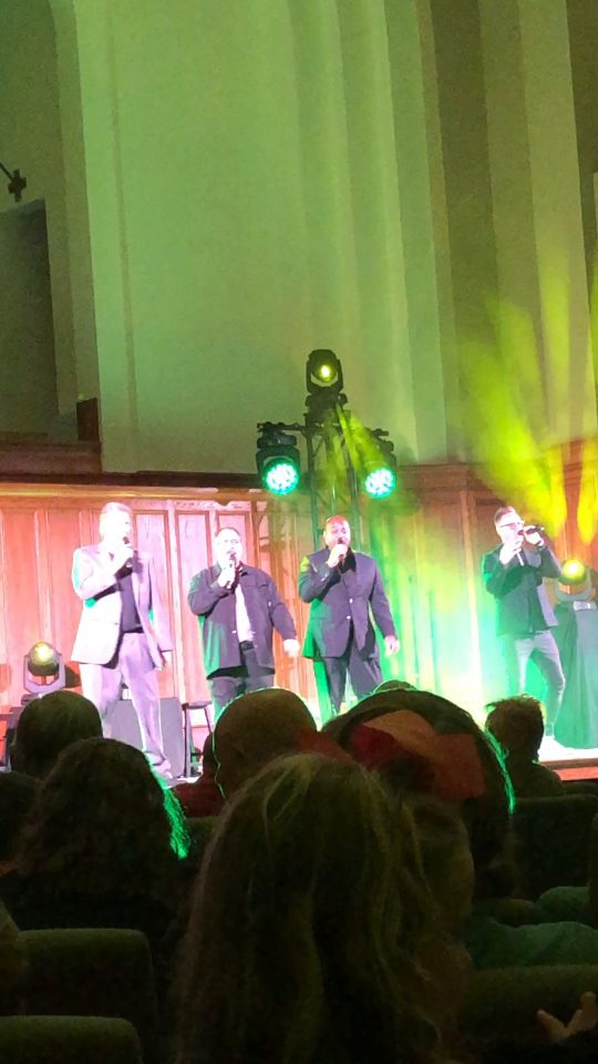 Acappella performing on stage during a concert, with four singers visible in front of an audience.
