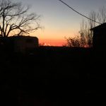 Sunset glowing orange beneath a fading sky, seen beyond dark silhouettes of trees and buildings.