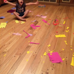 A young girl sits smiling in the middle of colorful paper scraps scattered across the floor during a craft project.
