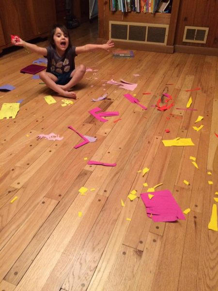 A young girl sits smiling in the middle of colorful paper scraps scattered across the floor during a craft project.