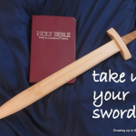 A wooden sword lies diagonally across a burgundy NIV Bible on dark blue fabric. White text beside it reads, “take up your sword!”