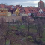 View of Rothenburg ob der Tauber, Germany, with red-roofed buildings, a church tower, medieval stone walls, and trees in the foreground.